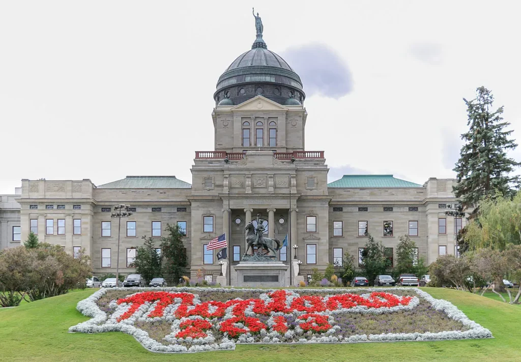 правото да изчисляваш „Montana State Capitol в Хелена, фасада с купол и цветна леха с надпис ‘Montana’ отпред“.​
